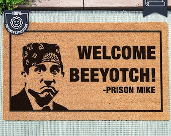May include: A brown doormat with the text "WELCOME BEEYOTCH! -PRISON MIKE" in black lettering. A black and white illustration of a man with a red bandana on his head is on the left side of the mat.
