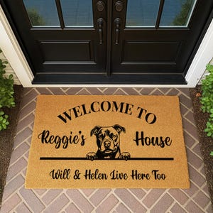 May include: A rectangular doormat with the text "WELCOME TO Reggie's House" in black. A black and white dog graphic is in the center. Below the dog is the text "Will & Helen Live Here Too". The mat is on a brick surface.