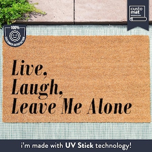 Op de afbeelding: Een bruine kokosvezel deurmat met de tekst "Live, Laugh, Leave Me Alone" in zwarte letters. De mat ligt op een blauw-wit gepatroneerd oppervlak. De tekst "i'm made with UV Stick technology!" staat onder de mat.