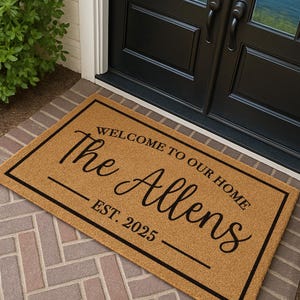 Puede incluir: Felpudo rectangular con borde negro y texto "WELCOME TO OUR HOME The Allens EST. 2025" en una fuente elegante. La alfombra es de color beige y est&aacute; sobre una superficie de ladrillo.
