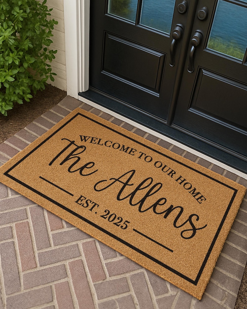 Puede incluir: Felpudo rectangular con borde negro y texto "WELCOME TO OUR HOME The Allens EST. 2025" en una fuente elegante. La alfombra es de color beige y est&aacute; sobre una superficie de ladrillo.