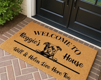 May include: A rectangular doormat with a tan background and black text. The text reads "WELCOME TO HOUSE" with "Reggie's" and "Will & Helen Live Here Too" in a script font. A black graphic of a dog is in the center.