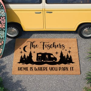 May include: A rectangular doormat with the text "The Fischers" and "HOME IS WHERE YOU PARK IT". The mat features a black camper silhouette, trees, a crescent moon, and stars. The mat is made of a natural fibre material.