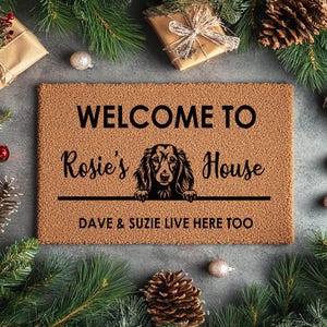 K&ouml;nnte beinhalten: Eine rechteckige Fu&szlig;matte mit dem Text "WELCOME TO Rosie's House" und "DAVE & SUZIE LIVE HERE TOO". Eine Hundeillustration befindet sich in der Mitte. Die Matte ist von Weihnachtsdekorationen umgeben, darunter Tannennadeln, Kugeln und verpackte Geschenke.