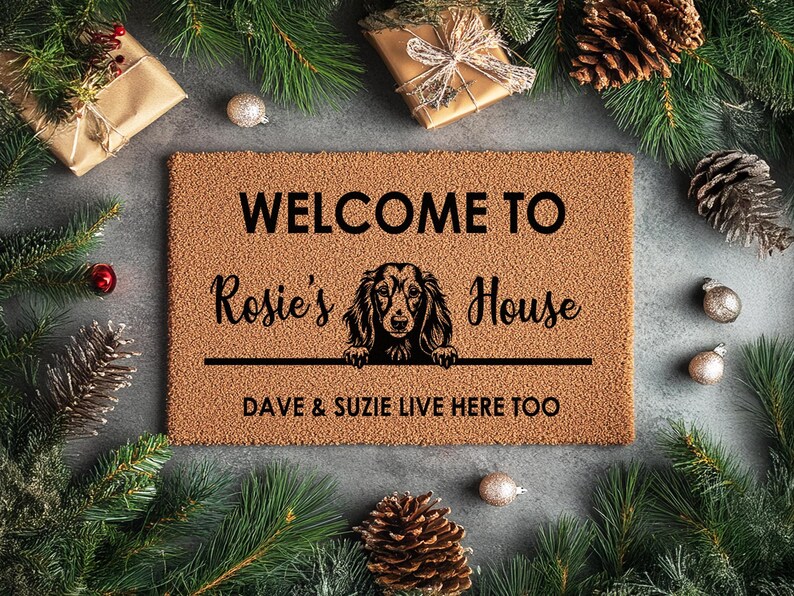 K&ouml;nnte beinhalten: Eine rechteckige Fu&szlig;matte mit dem Text "WELCOME TO Rosie's House" und "DAVE & SUZIE LIVE HERE TOO". Eine Hundeillustration befindet sich in der Mitte. Die Matte ist von Weihnachtsdekorationen umgeben, darunter Tannennadeln, Kugeln und verpackte Geschenke.