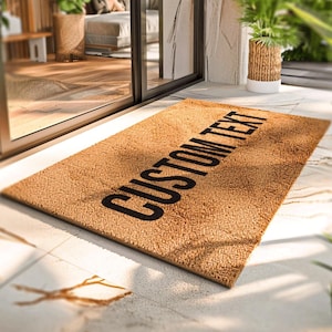 Pode incluir: Um capacho retangular feito de fibra de coco castanha natural. O tapete apresenta as palavras "CUSTOM TEXT" em letras pretas em negrito no centro. O tapete é colocado sobre uma superfície de azulejos perto de uma porta de vidro.