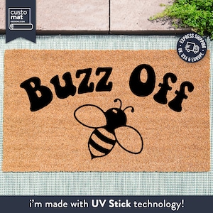 May include: A brown coir doormat with the text "Buzz Off" in black stylized lettering. A black and white cartoon bee is centred below the text. The doormat is made with UV Stick technology.