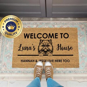 May include: A rectangular doormat with the text "WELCOME TO Luna's House" and a Persian cat illustration. Below the text is "HANNAH & JESSE LIVE HERE TOO". The mat is on a patterned rug.