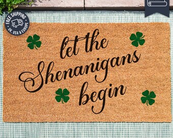 Puede incluir: Una alfombra de coco marrón con el texto "Let the Shenanigans Begin" en letras negras cursivas. Cuatro diseños de trébol de cuatro hojas verdes están dispersos alrededor del texto.