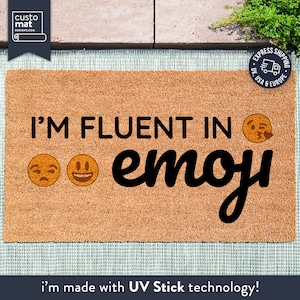 May include: A brown coir doormat with the text "I'm fluent in emoji" in black lettering. The text is accompanied by three yellow emoji faces. The doormat is made with UV Stick technology.