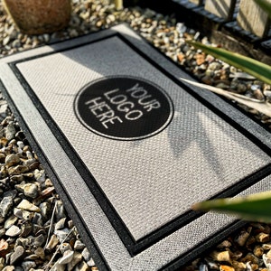 Puede incluir: Una alfombra de bienvenida gris con un borde negro y un c&iacute;rculo negro en el centro que dice "YOUR LOGO HERE".