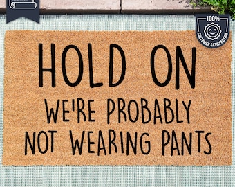 May include: A brown doormat with the text "HOLD ON WE'RE PROBABLY NOT WEARING PANTS" in black lettering. The doormat is made with UV Stick technology.