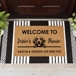 K&ouml;nnte beinhalten: Eine rechteckige Fu&szlig;matte mit dem Text "WELCOME TO Jessie's House" und einer Hunde-Grafik. Die Matte ist braun mit schwarzem Text und einem schwarz-wei&szlig; gestreiften Rand. Der Text "KAITLYN & GEORGE LIVE HERE TOO" ist ebenfalls enthalten.