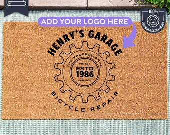 May include: A brown coir doormat with the text "ADD YOUR LOGO HERE" and "HENRY'S GARAGE" in black. The text "THE PROFESSIONAL FINEST ESTD 1986 SERVICE" is inside a black gear with the text "BICYCLE REPAIR" below. A 100% customer satisfaction guarantee badge is in the top right corner.