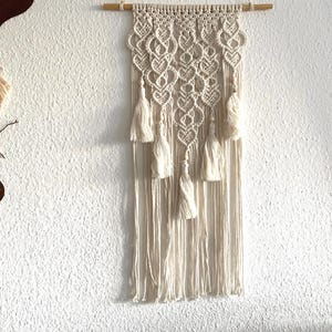 Könnte beinhalten: Cremefarbener Makramee-Wandbehang mit einem Holzstab. Das Design weist komplizierte Knotenmuster und hängende Quasten auf. Dieses handgefertigte Stück verleiht jedem Raum einen Hauch von Bohème. Die Wanddekoration wird von einer Naturfaserschnur gehalten.