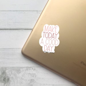 Puede incluir: Una pegatina blanca con la frase "MAKE TODAY A GOOD DAY" en letras rojas, adherida a un iPad dorado. La pegatina tiene forma de nube. El iPad está sobre una superficie de madera clara.