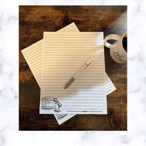May include: Two sheets of lined paper with a drawing of a stack of books and a lamp on the bottom sheet. A pen is lying on top of the paper. The paper is on a dark brown wooden surface.
