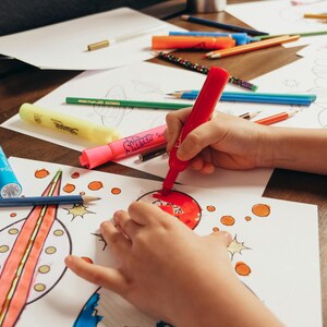 May include: A child's hands coloring a drawing with a red marker. The image shows various colored pencils, markers, and drawings on a wooden surface, suggesting a creative activity. The drawings feature a rocket ship and planets.