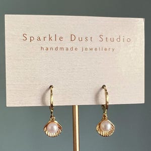 May include: Gold-coloured hoop earrings, each featuring a seashell-shaped charm with a single, round, white pearl. The earrings are displayed on a card with the text "Sparkle Dust Studio handmade jewellery".