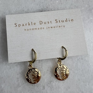 May include: Gold-tone hoop earrings with a celestial design. Each earring has a small, circular charm featuring a sun and moon motif. The earrings are presented on a white card with the text "Sparkle Dust Studio handmade jewellery".
