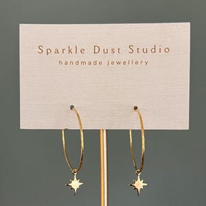 May include: Gold hoop earrings with a star-shaped charm. The earrings are presented on a white card with the text "Sparkle Dust Studio handmade jewellery". The earrings are suspended from a gold stand.