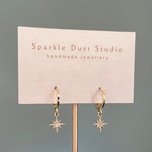 May include: Gold-coloured hoop earrings with starburst charms. The earrings are displayed on a card that reads "Sparkle Dust Studio handmade jewellery."