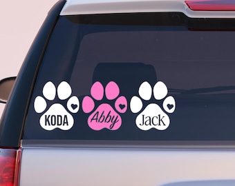 May include: Three paw print car decals. Two white paw prints with the names "Koda" and "Jack" and a pink paw print with the name "Abby". Each paw print has a heart symbol.