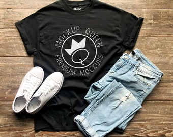 Puede incluir: Una camiseta negra con un diseño circular blanco que dice "Mockup Queen Premium Mockups" con una corona y una letra Q dentro del círculo. La camiseta está colocada sobre una superficie de madera con un par de zapatillas blancas y un par de pantalones vaqueros azules.