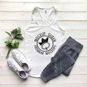 May include: White tank top with a black circle logo that says "Mockup Queen Premium Mockups" with a crown and a letter Q inside the circle. The tank top is laying on a white wooden surface with a pair of white sneakers and gray leggings.