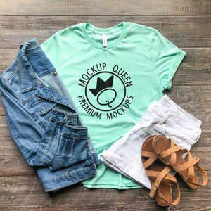 May include: A mint green t-shirt with a black circle graphic that says "Premium Mockups Queen" with a crown above the Q. The shirt is lying on a wooden surface with a denim jacket, white jeans, and brown sandals.
