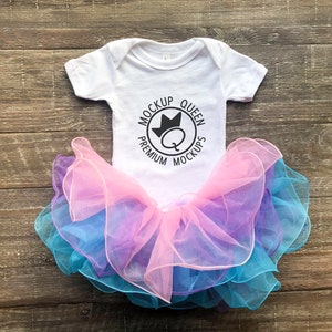 May include: White baby onesie with short sleeves and a black circle graphic that says "Mockup Queen Premium Mockups" with a crown above the Q. The onesie is on top of a multi-layered tulle skirt in shades of pink, purple, and blue.