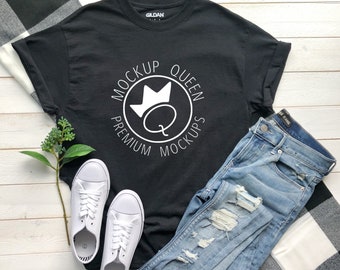 May include: Black t-shirt with a white circle design that says "Mockup Queen Premium Mockups" with a crown and a letter Q inside the circle. The shirt is laid flat on a white wooden surface with a pair of white sneakers, blue jeans, and a black and white plaid blanket.