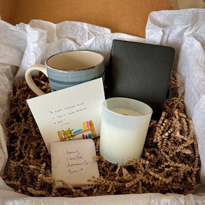 May include: A gift box containing a blue striped ceramic mug, a black notebook, a white candle in a frosted glass jar, and a card with the text A room without books is like a body without a soul - Cicero, plus a small packet labeled honey vanilla chamomile tea.