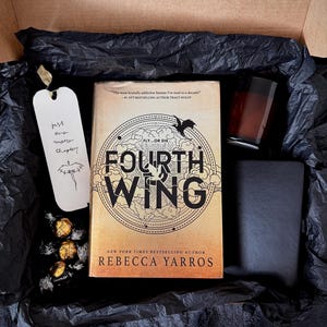 May include: A gift box containing the book Fourth Wing by Rebecca Yarros, a white bookmark with the text 'just one more chapter' and a dragon illustration, a dark candle in a glass jar, a black notebook, and three gold-wrapped chocolate truffles.