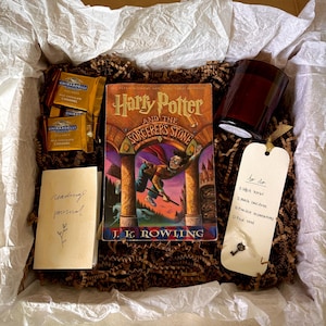 May include: A gift box containing a copy of Harry Potter and the Sorcerer's Stone, two Ghirardelli milk chocolate caramel squares, a dark glass candle, a small reading journal, and a white tag with a key and a handwritten to-do list.