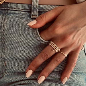 Könnte beinhalten: Ein Satz von vier goldenen Ringen mit weißen Perlen und einem kleinen Sonnenanhänger. Die Ringe sind an einem Finger übereinander getragen.