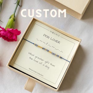 May include: A blue and gold beaded bracelet with the words "DOG LOVER" spelled out in Morse code on a white card in a beige box. The card also reads "What greater gift than the love of a dog." and "Morse Code Collection."