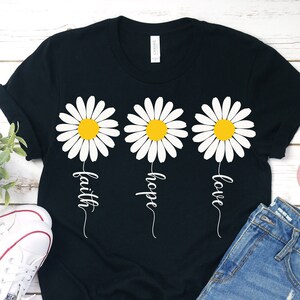 May include: Black t-shirt with three white daisies and the words "faith", "hope", and "love" written underneath each daisy.