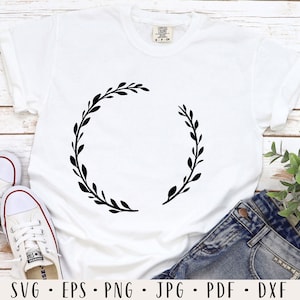 Puede incluir: Una camiseta blanca con un dise&ntilde;o de corona floral negra. La corona es un c&iacute;rculo simple con hojas y ramas.