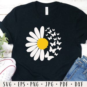 May include: Black t-shirt with a white daisy and yellow center surrounded by white butterflies. The shirt is on a wooden surface with a pair of white sneakers and blue jeans.