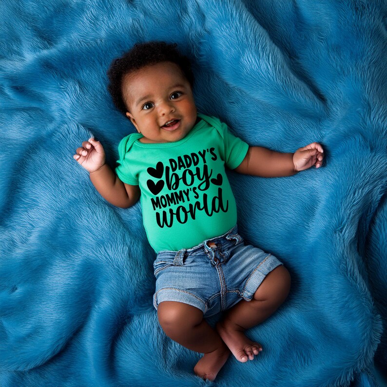 Pu&ograve; includere: Un bambino che indossa un body verde con il testo "Daddy's Boy Mommy's World" in lettere nere e tre cuori neri. Il bambino &egrave; sdraiato su una coperta blu pelosa.