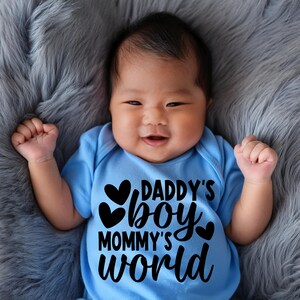 Pu&ograve; includere: Un bambino che indossa un body azzurro chiaro con testo nero che recita "Daddy's Boy Mommy's World" e tre cuori neri.