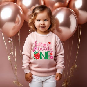 Puede incluir: Una sudadera rosa con el texto "berry sweet one" en letras verdes y rosas. Una fresa roja con hojas verdes est&aacute; al lado de la palabra "one".