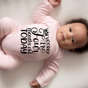 May include: A pink baby onesie with black text that reads 'HOORAY I'M Four months old TODAY' with a crown above the word 'HOORAY'.