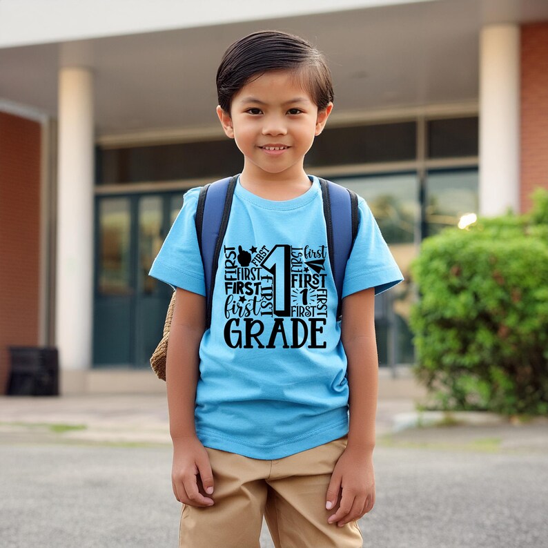 May include: A young boy wearing a light blue t-shirt with a black graphic that says "First Grade" with the number "1" surrounded by the word "First" in various sizes and orientations. He is standing in front of a school building and wearing a backpack.
