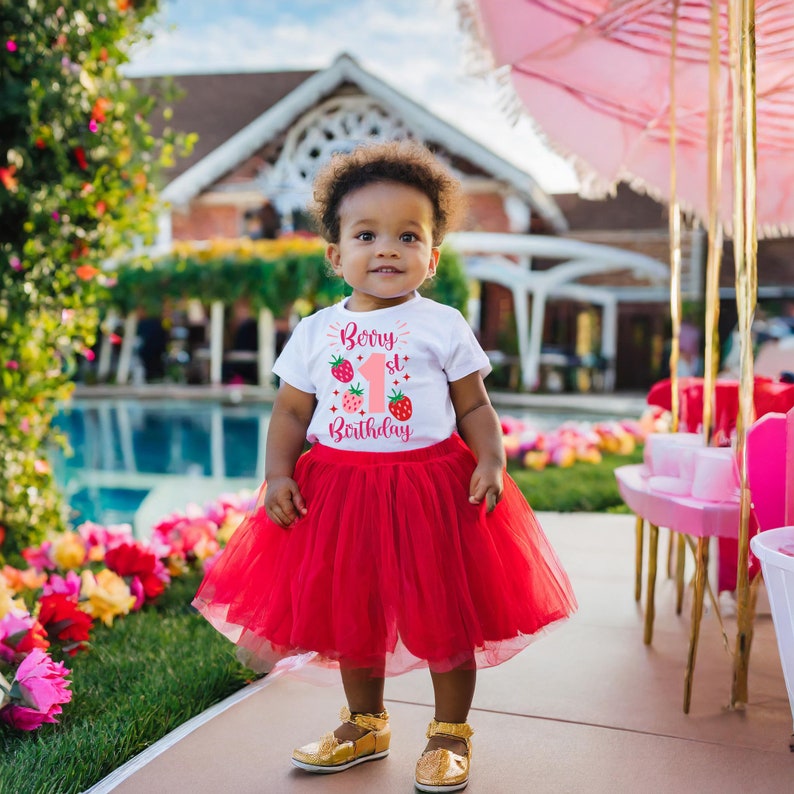 May include: A white t-shirt with a pink and red graphic that says "Berry 1st Birthday" with strawberries. A child is wearing the shirt and a red tulle skirt.