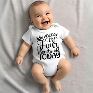 May include: A baby wearing a white bodysuit with black text that reads 'Hooray I'm Four months old TODAY' with a crown above the word 'Hooray'.