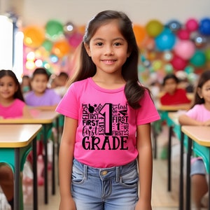 May include: A young girl wearing a pink t-shirt with the words "First Grade" printed in black. The shirt features the number "1" surrounded by the word "first" repeated multiple times. The girl is standing in a classroom setting.
