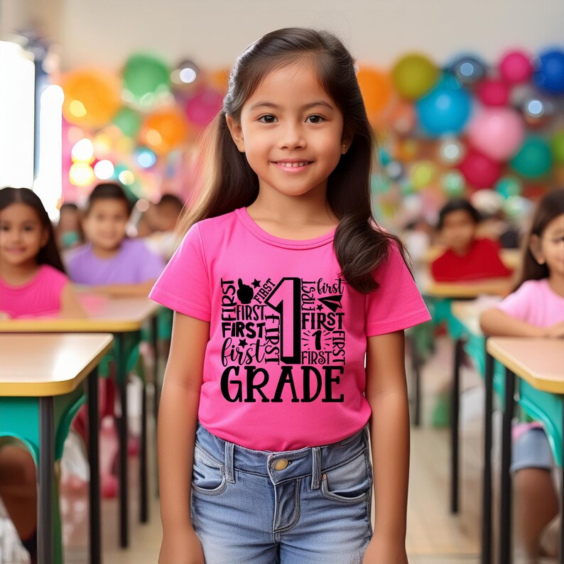 May include: A young girl wearing a pink t-shirt with the words "First Grade" printed in black. The shirt features the number "1" surrounded by the word "first" repeated multiple times. The girl is standing in a classroom setting.