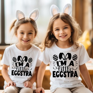 May include: Two children wearing white t-shirts with black bunny ears and the text "I'm a little eggstra" printed on them. The children are smiling and looking at the camera.
